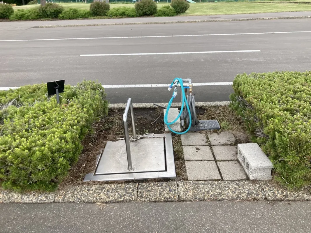 てんとらんどオートキャンプ場：植え込みの間の金属のふたと、ホースのついた水道